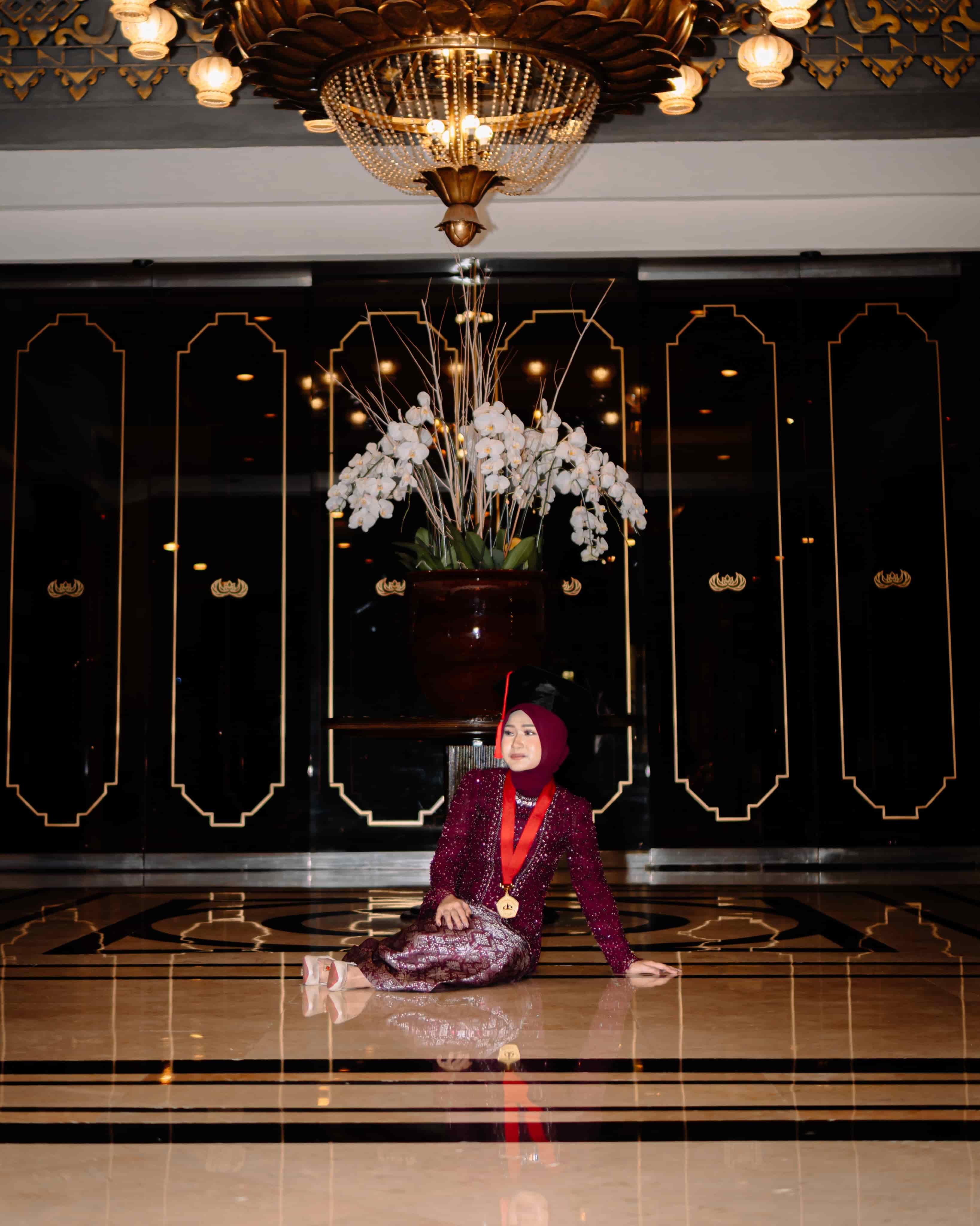 Law graduate in a maroon kebaya, gown, and alumni medal seated in a ballroom under a chandelier with white orchids