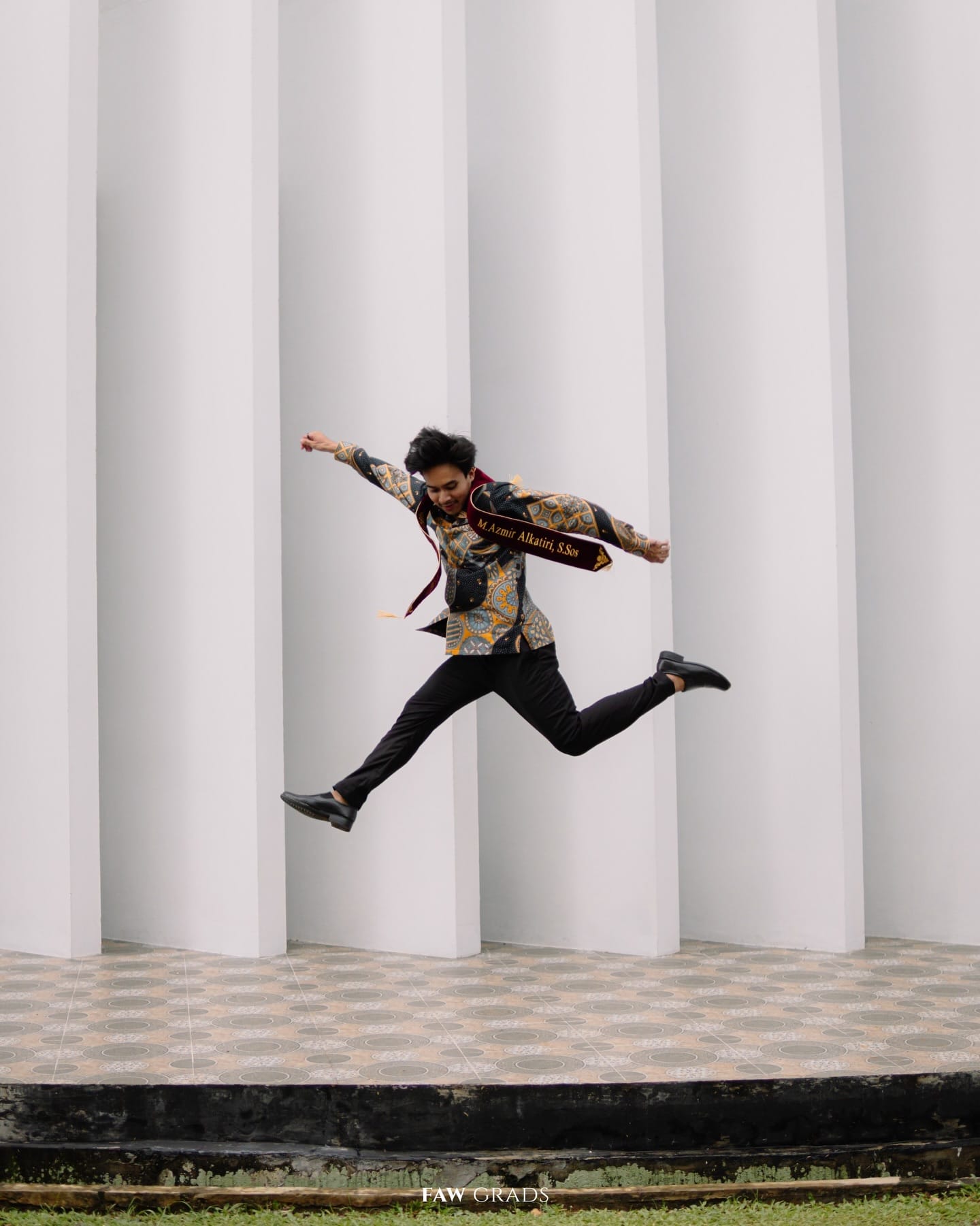 Graduate in a dark batik shirt with a maroon sash jumping in front of the white pillars of UIN Jakarta