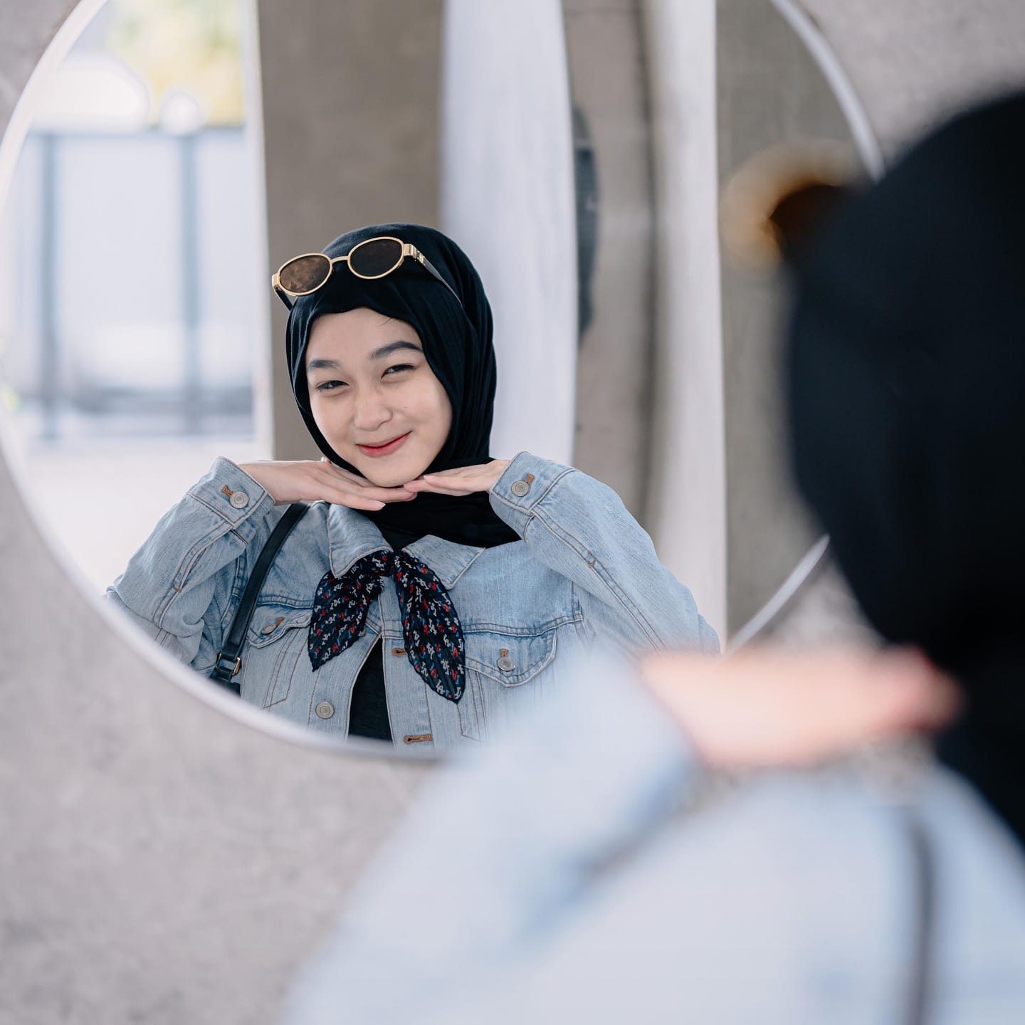 Potret pantulan di cermin bundar: perempuan berhijab hitam berjaket denim tersenyum ke cermin