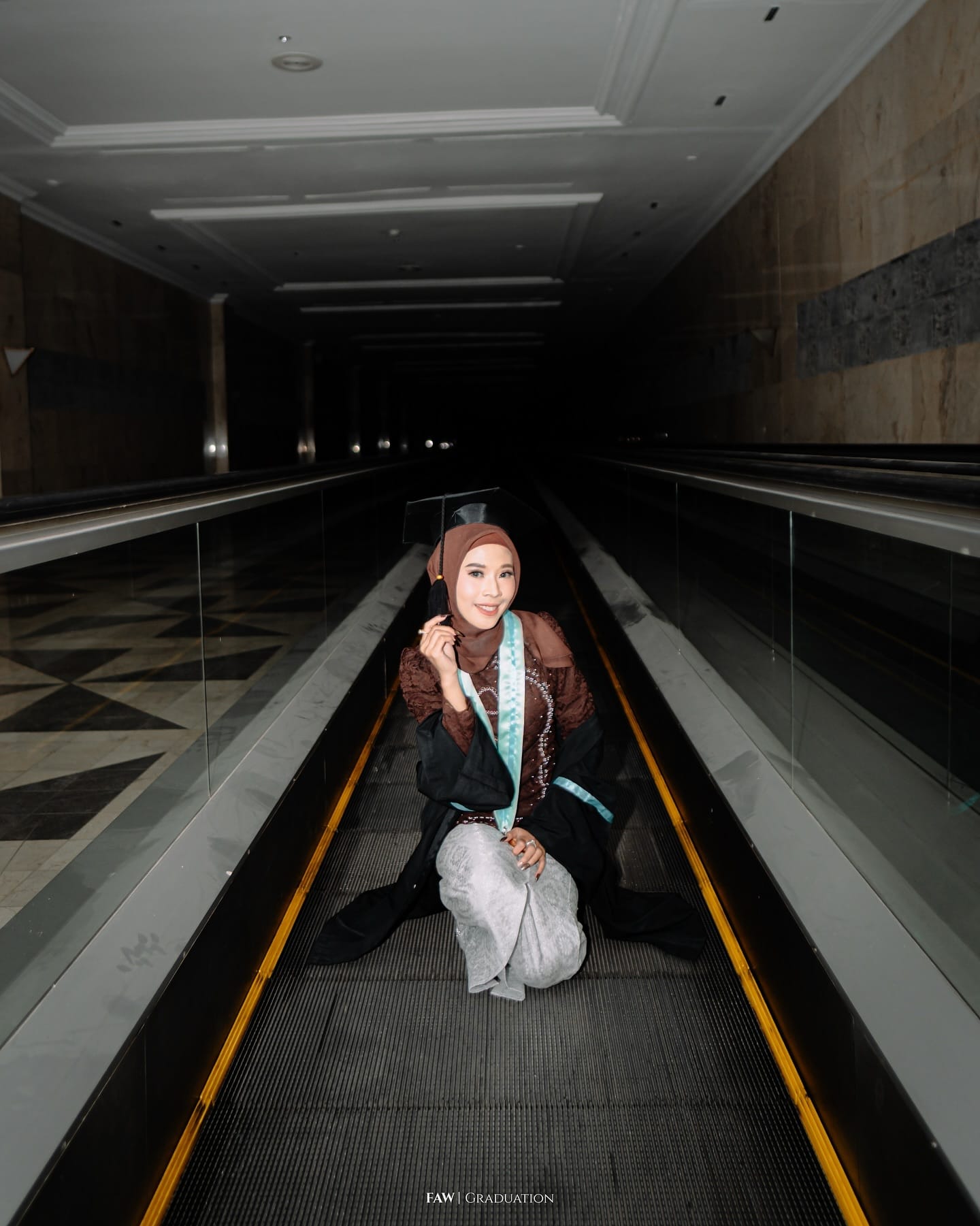 Wisudawati duduk di travelator dengan toga pada sesi malam, garis kuning eskalatornya menyala