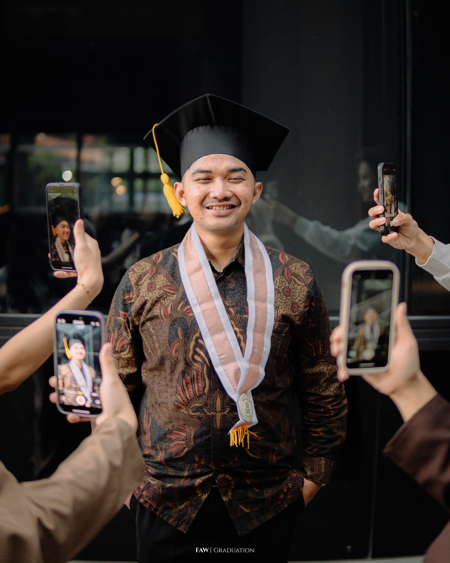 Wisudawan berkemeja batik dan toga dikelilingi tangan teman-teman yang memotretnya dengan ponsel