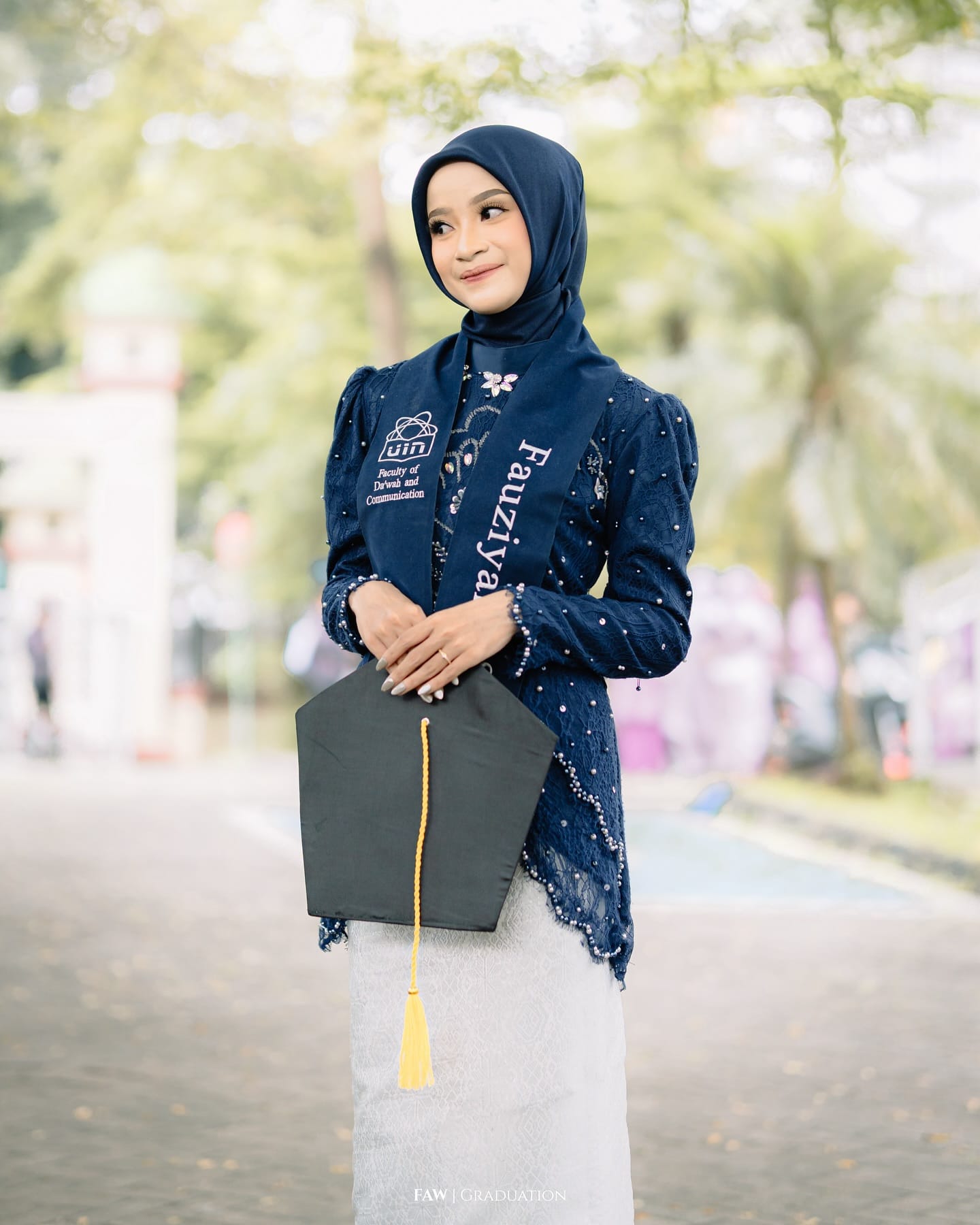 Wisudawati berkebaya biru dongker memegang toga, selempang Faculty of Dakwah and Communication UIN Jakarta