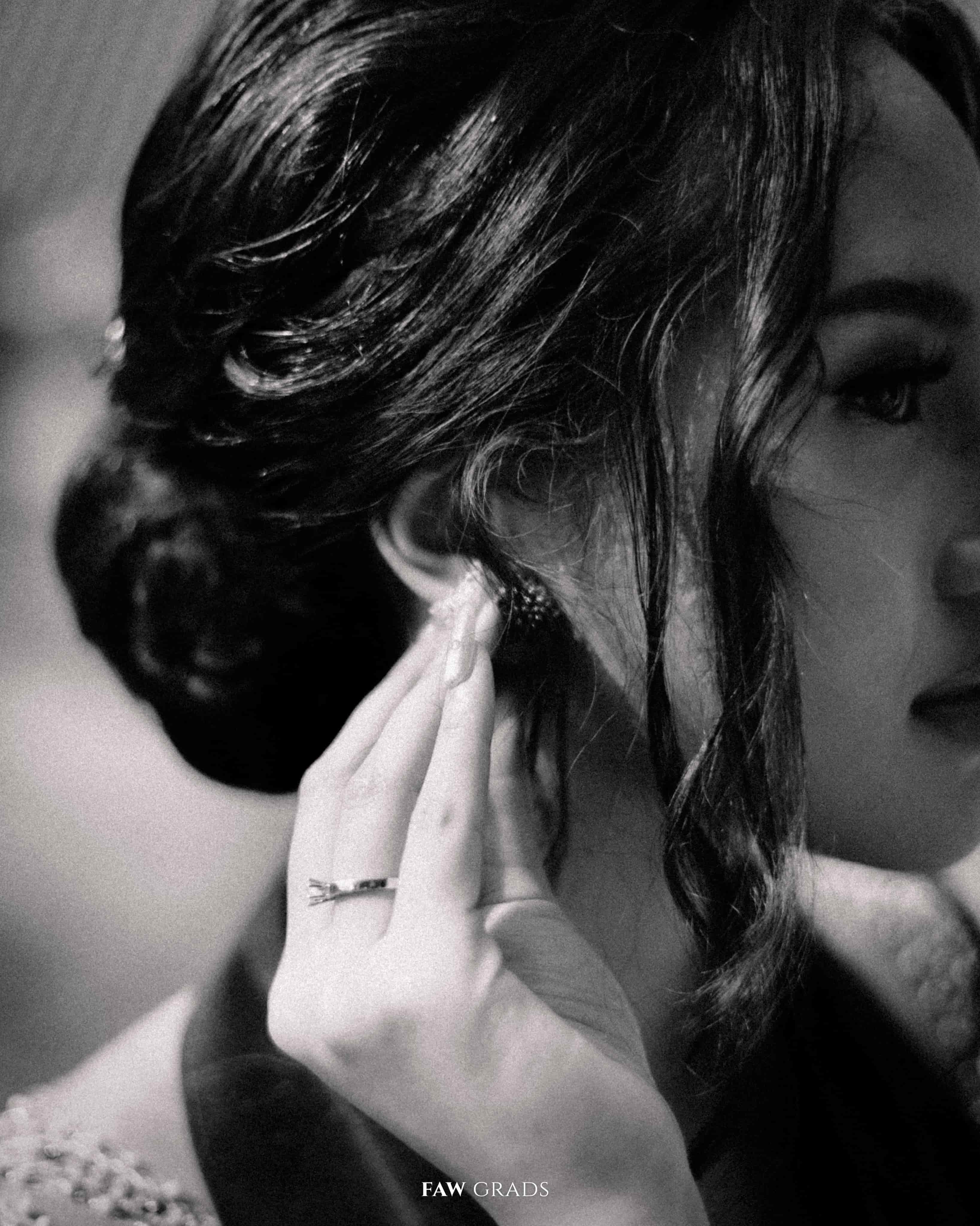 Foto close-up hitam putih bergrain wisudawati Universitas Indonesia menyentuh anting, helai rambut lepas menutupi wajah, kebaya berpayet di bawah bingkai
