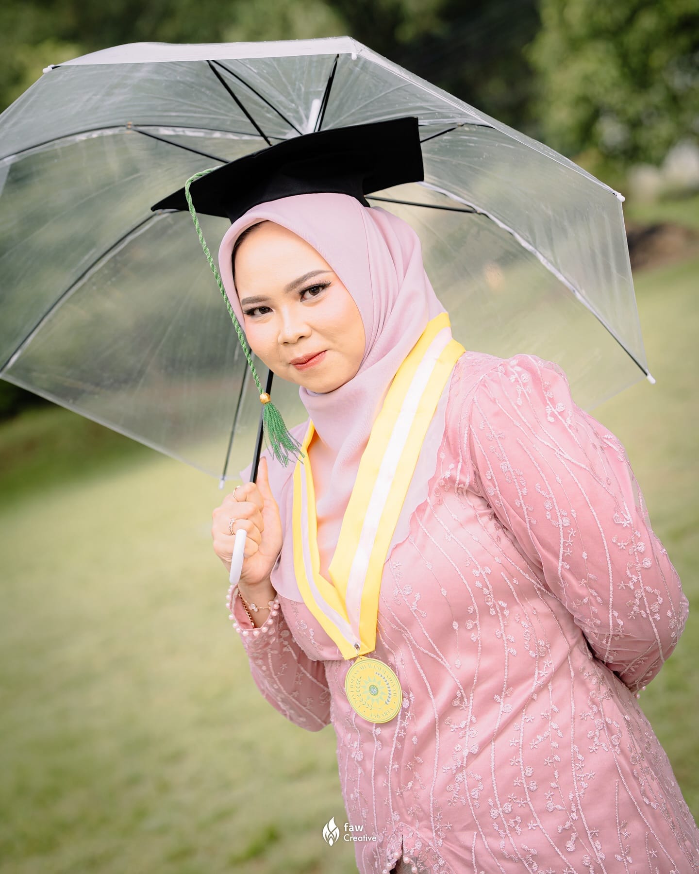 Wisudawati berkebaya merah muda berpayung bening saat gerimis, medali Universitas Muhammadiyah Jakarta di dada