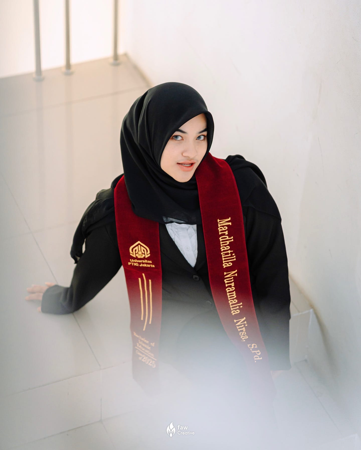 Wisudawati Universitas PTIQ Jakarta menoleh ke kamera dari tangga, selempang maroon bertuliskan namanya