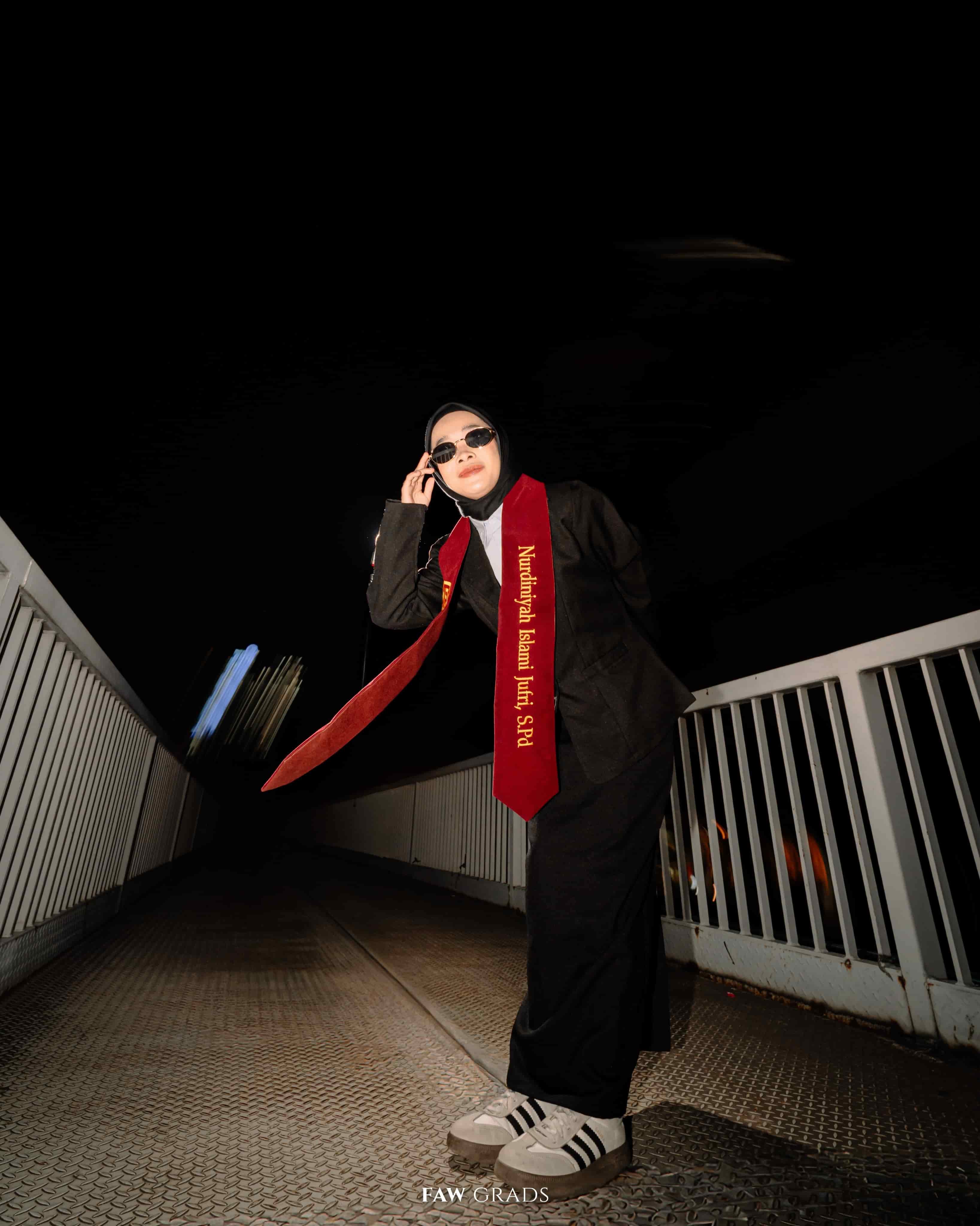 Foto wisuda malam di jembatan kota, wisudawati hijab memakai toga hitam, selempang merah, sunglasses, dan sneakers bergaya urban