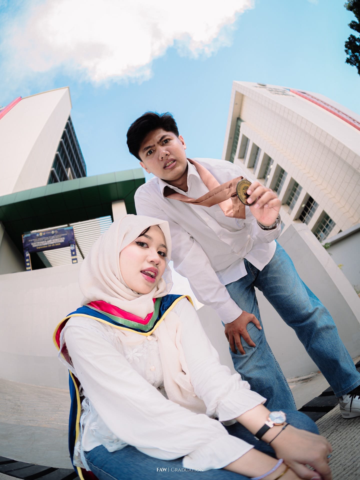 Dua sahabat bergaya di depan gedung kampus dengan medali dan selempang, diambil dengan lensa fisheye