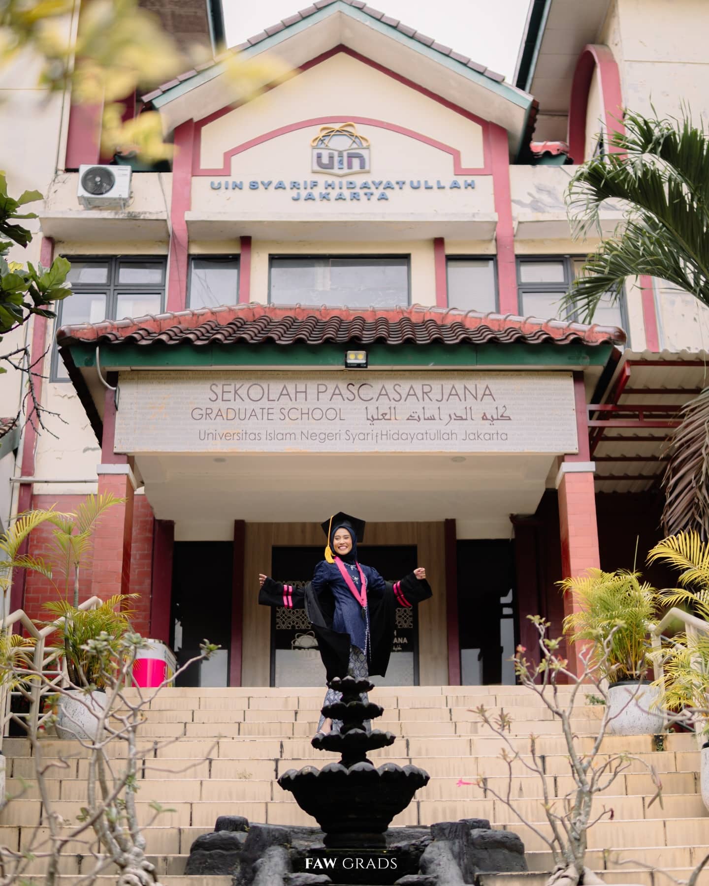 Graduate in hijab opening her arms in joy in front of the UIN Jakarta Graduate School building, black gown with a magenta hood